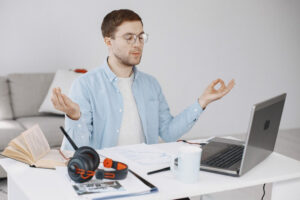A person wearing a light blue shirt sits at a desk, meditating with hands in a gesture, surrounded by books, headphones, and a laptop.