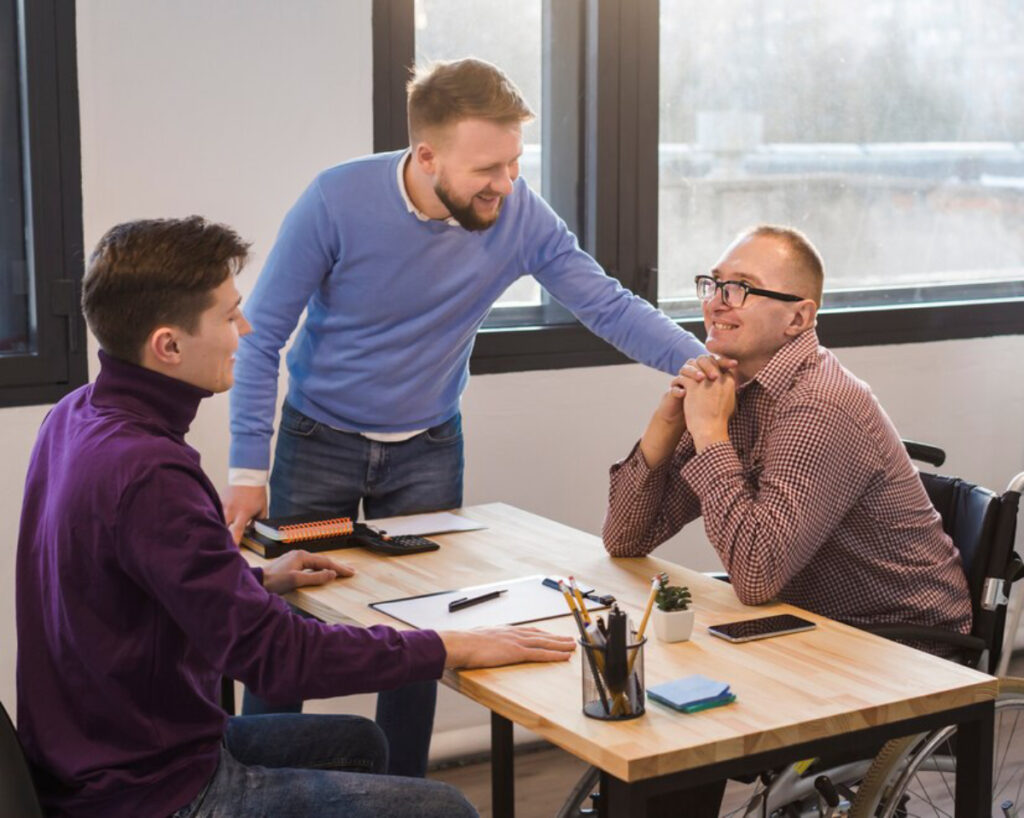 Three individuals engage in a discussion at a wooden table, with notebooks and pens, in a bright, modern office setting.
