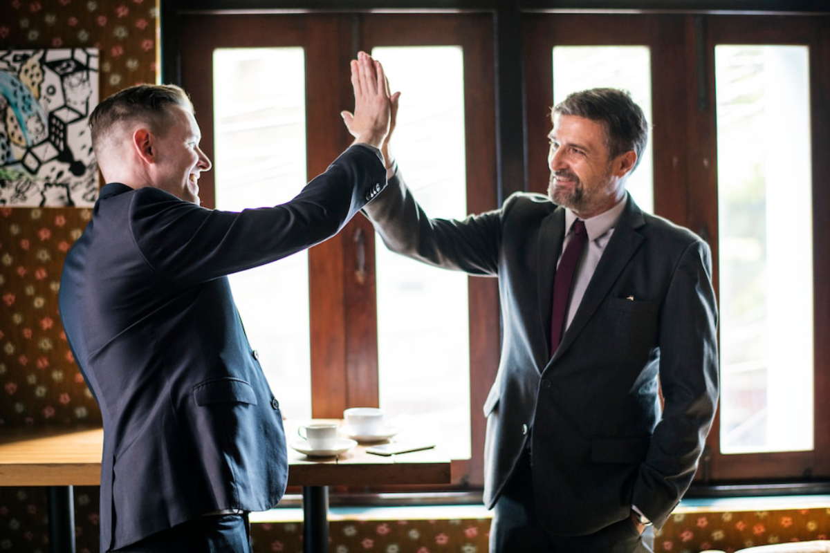 Two men in formal suits are giving each other a high-five inside a cafe with dark wooden windows and a patterned wall in the background.