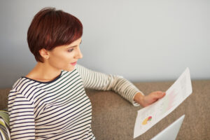 A woman in a striped shirt sits on a couch, reviewing a document with colorful graphs and charts. A laptop rests nearby.