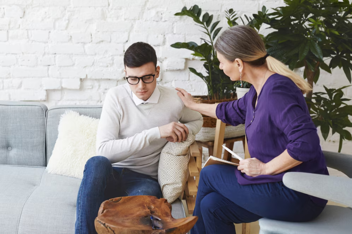 A woman in a purple top offers comfort to a man sitting on a gray sofa, surrounded by plants in a cozy, well-lit room.