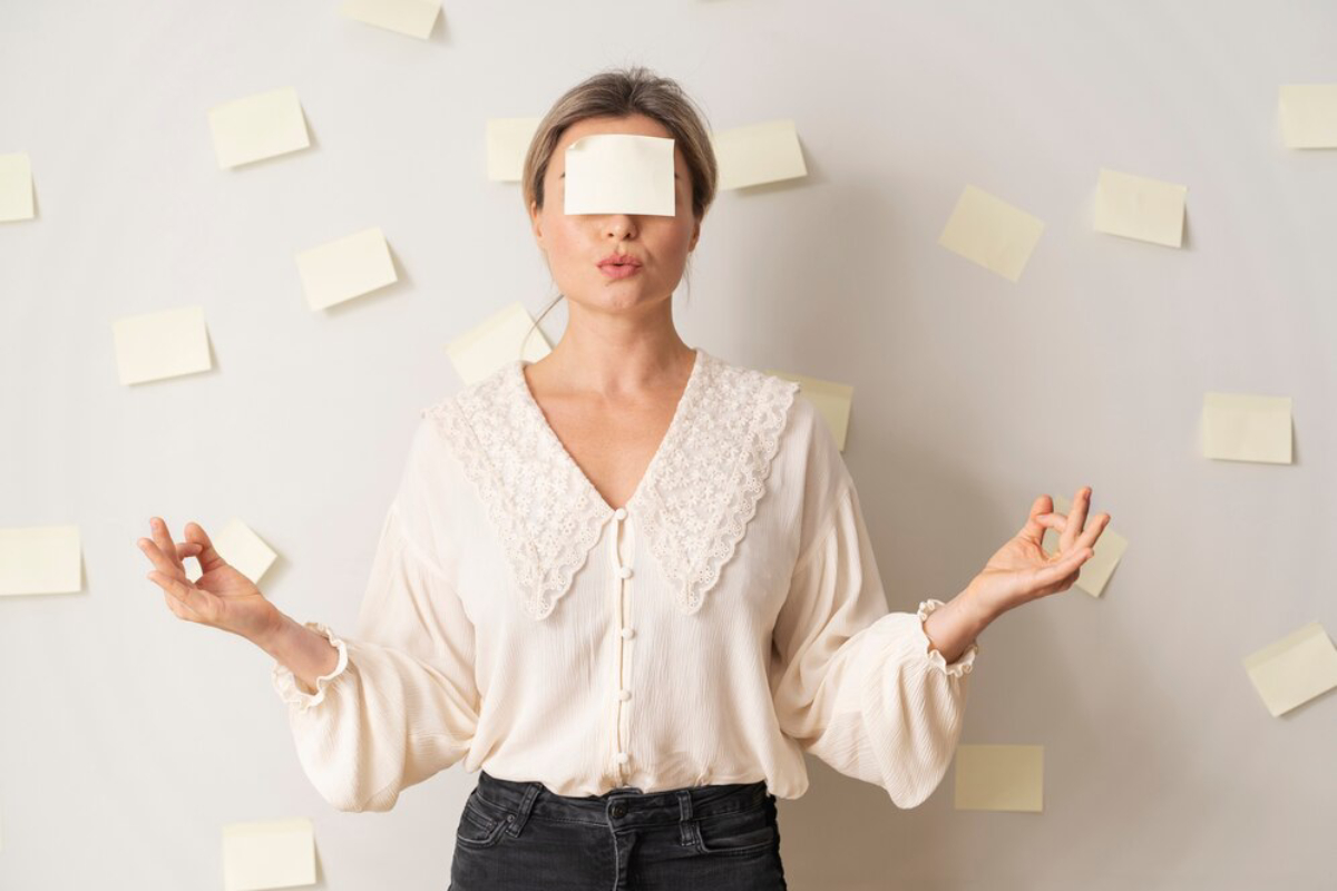 A person in a light-colored blouse is standing in a meditative pose in front of a wall adorned with pale sticky notes.