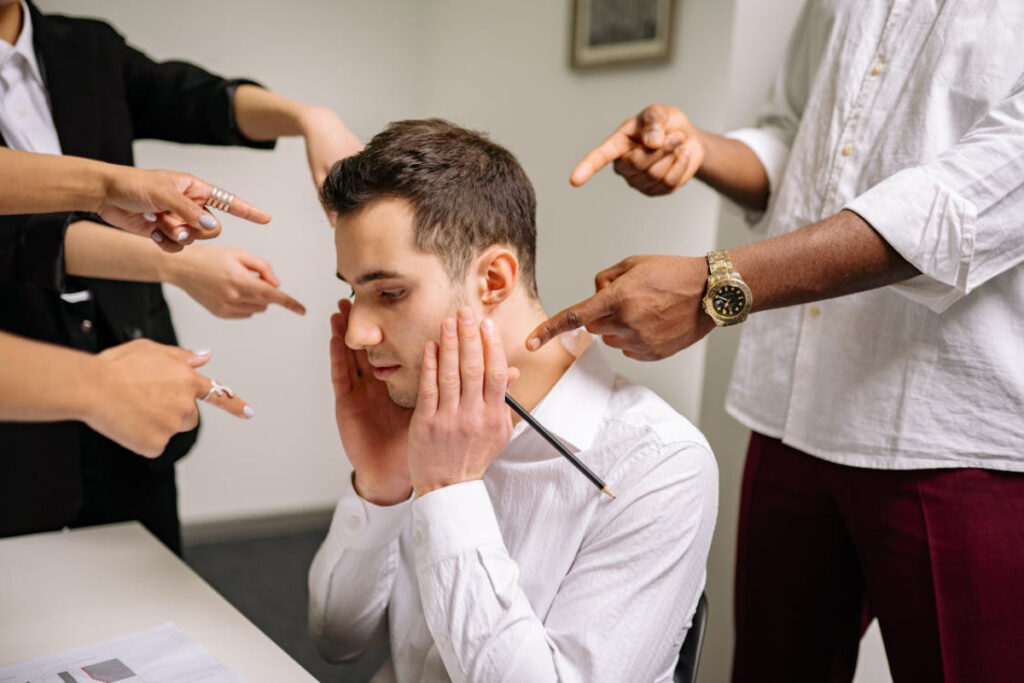 A group of people in formal attire point emphatically at a seated individual holding a pencil, expressing accusation or confrontation.