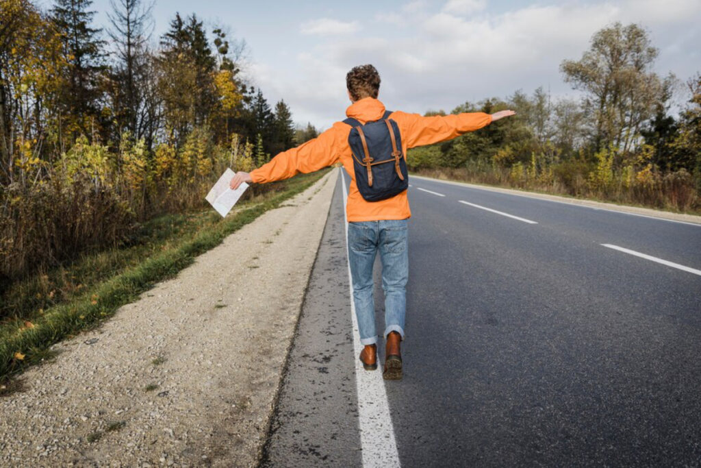 A person in an orange jacket and backpack walks along a roadside, arms outstretched, holding a map against a backdrop of autumn foliage.