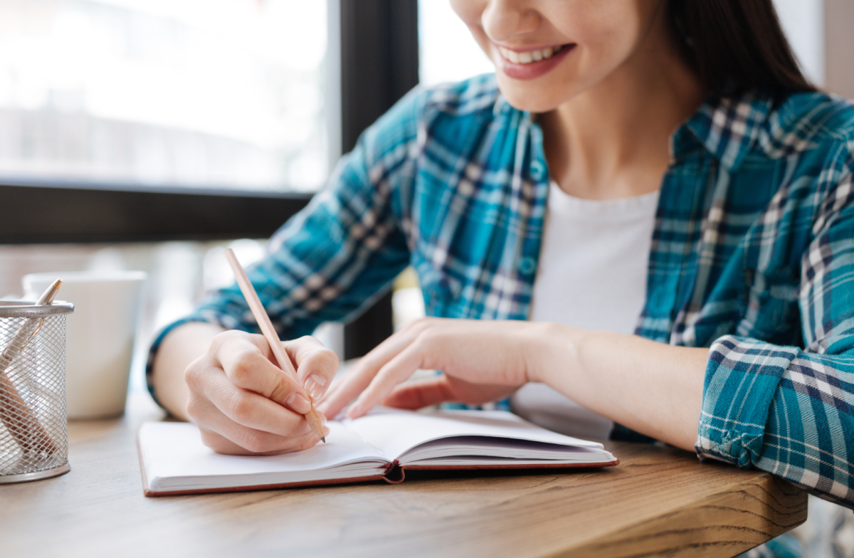 A young woman in a checkered shirt smiles as she writes in a notebook at a café table with a cup beside her.