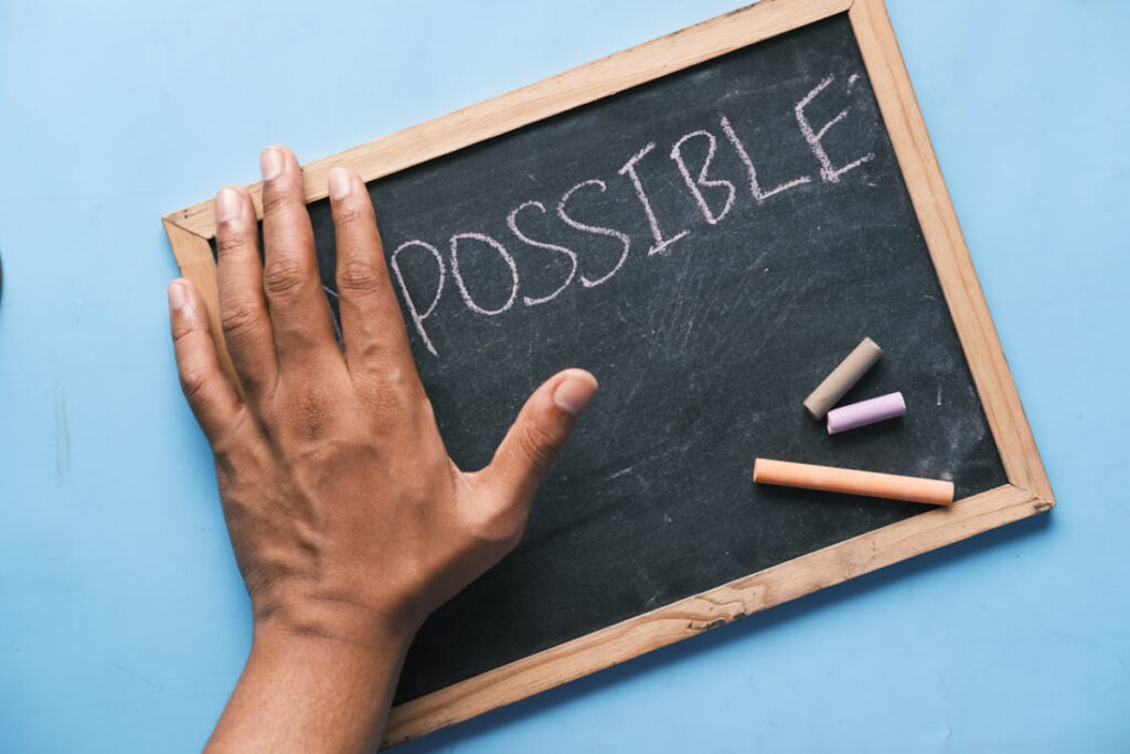 A hand resting on a chalkboard displaying the word POSSIBLE, with three pieces of colored chalk nearby on a blue background.