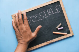A hand resting on a chalkboard displaying the word POSSIBLE, with three pieces of colored chalk nearby on a blue background.