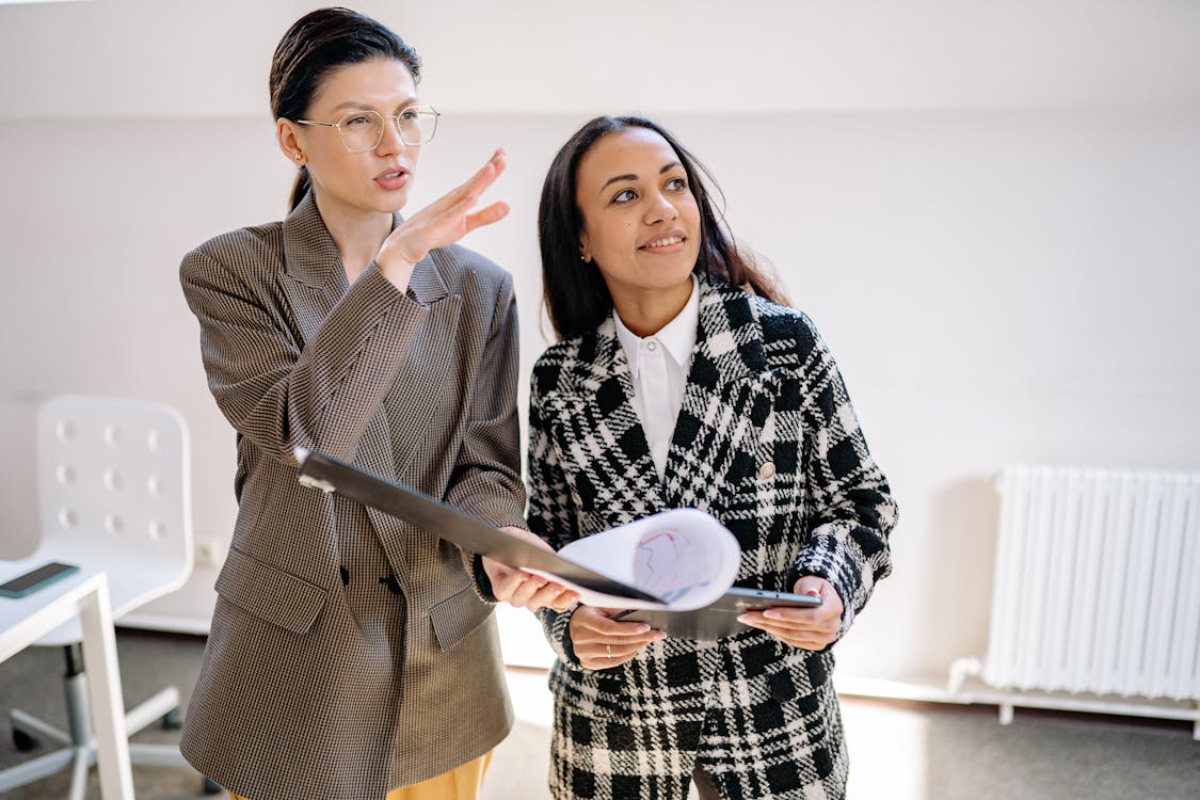 Two women in stylish outfits discuss a document while standing in a modern office space with minimalistic decor.