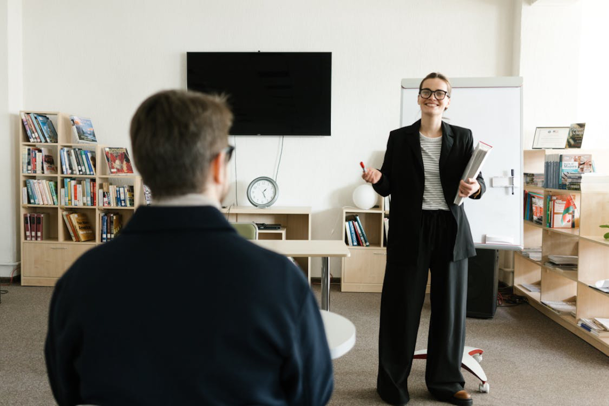 A person in a black suit stands gesturing while speaking to an audience in a modern classroom with bookshelves in the background.