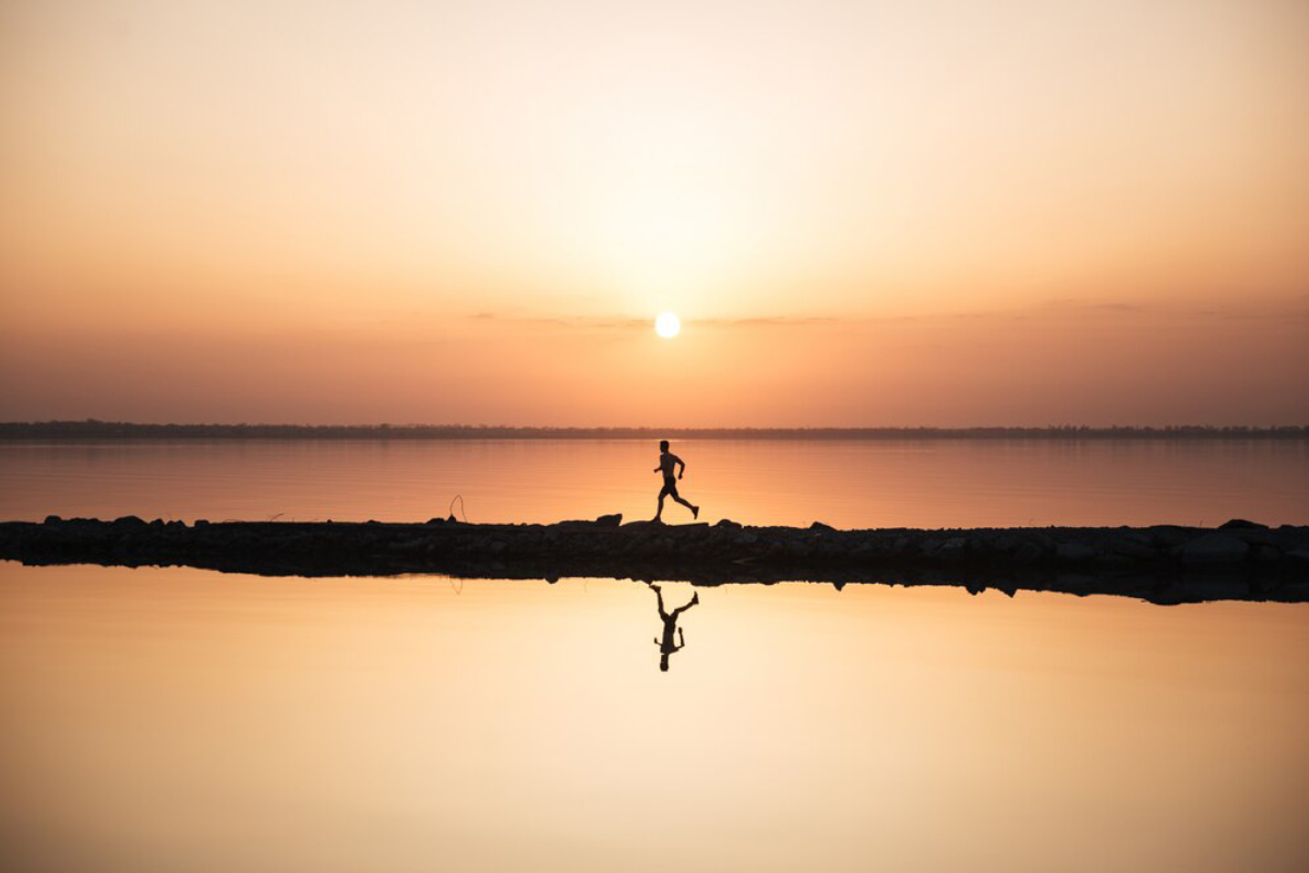 A silhouette of a runner on a rocky path by a calm lake at sunrise, reflecting golden hues in the water.