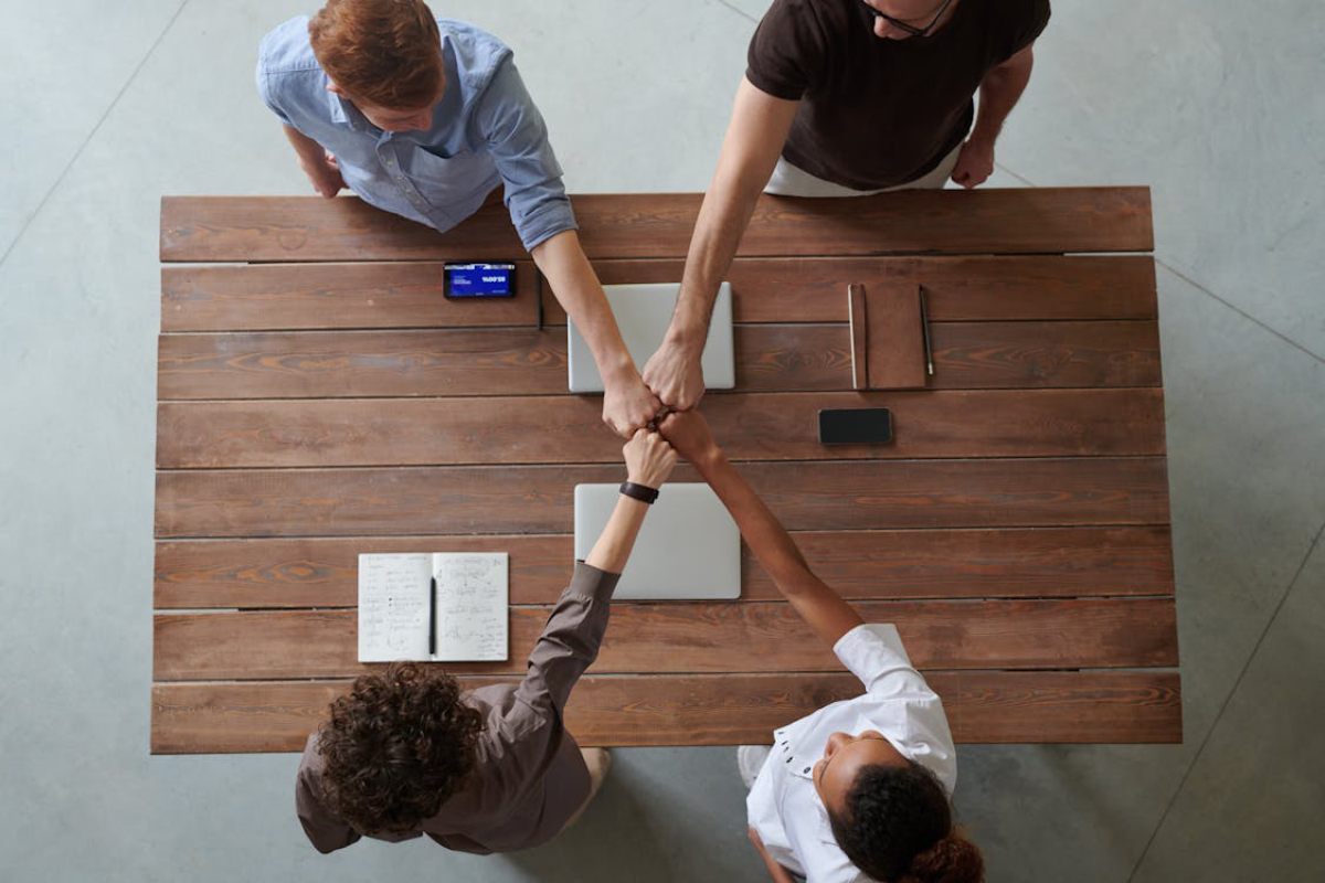 Four hands coming together in a fist bump over a wooden table, with a laptop, notebooks, and smartphones nearby.