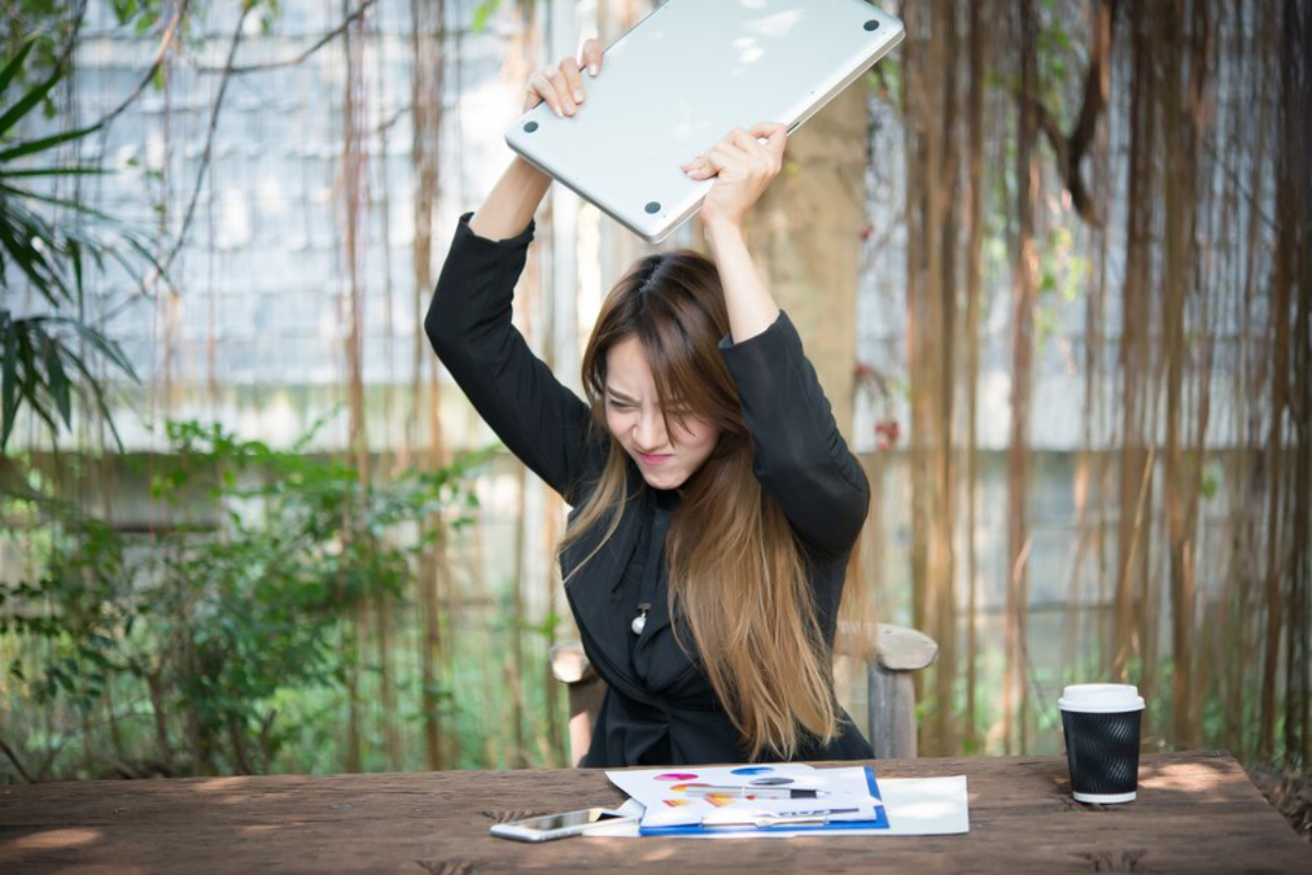 A person in a black jacket raises a laptop above their head in frustration or anger, amid a lush outdoor setting with a coffee cup on the table.