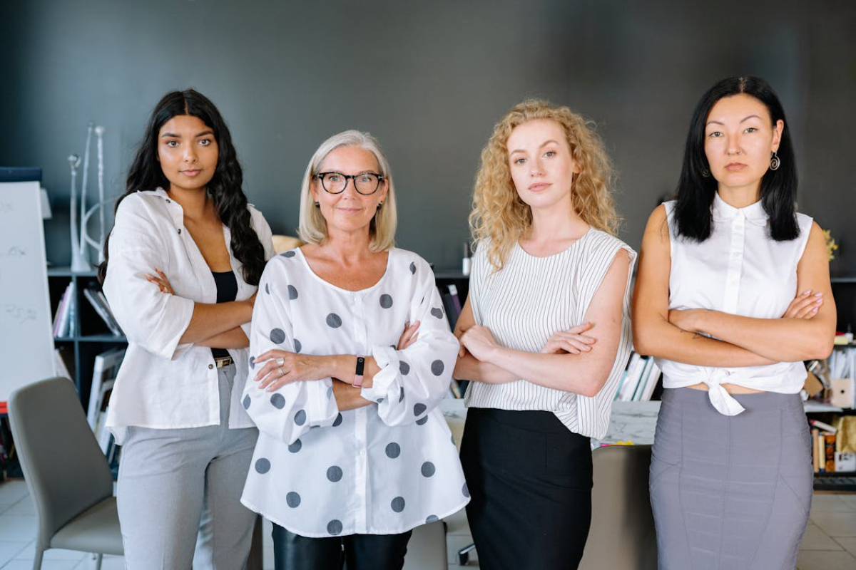 Four women stand confidently with arms crossed in a modern office setting, showcasing diverse styles and professional attire.