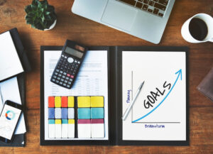 An organized workspace featuring a calculator, colorful sticky notes, a laptop, and a goal chart