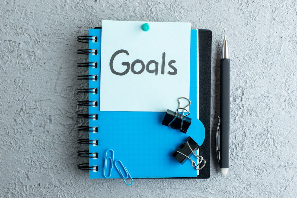 A blue notebook with a sticky note labeled Goals," accompanied by paper clips and a black pen on a gray textured surface.