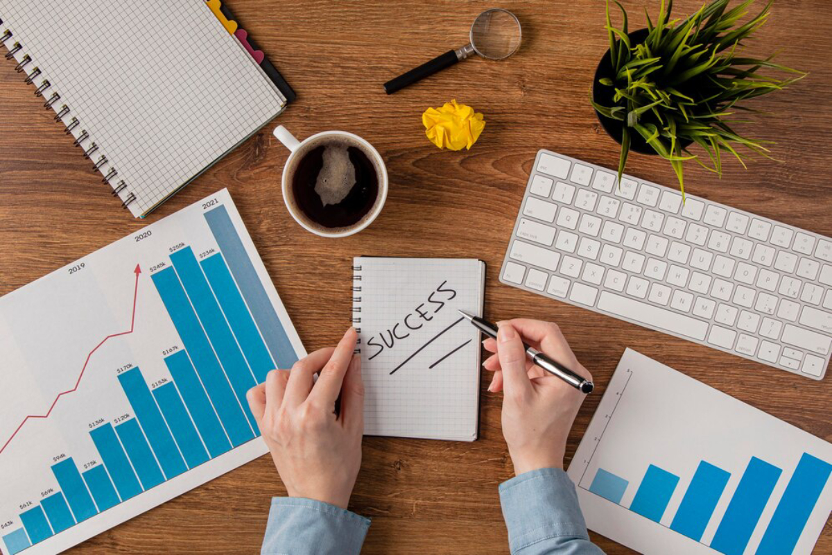 A person writes SUCCESS in a notebook while analyzing growth charts, a cup of coffee, and office supplies on a wooden desk.