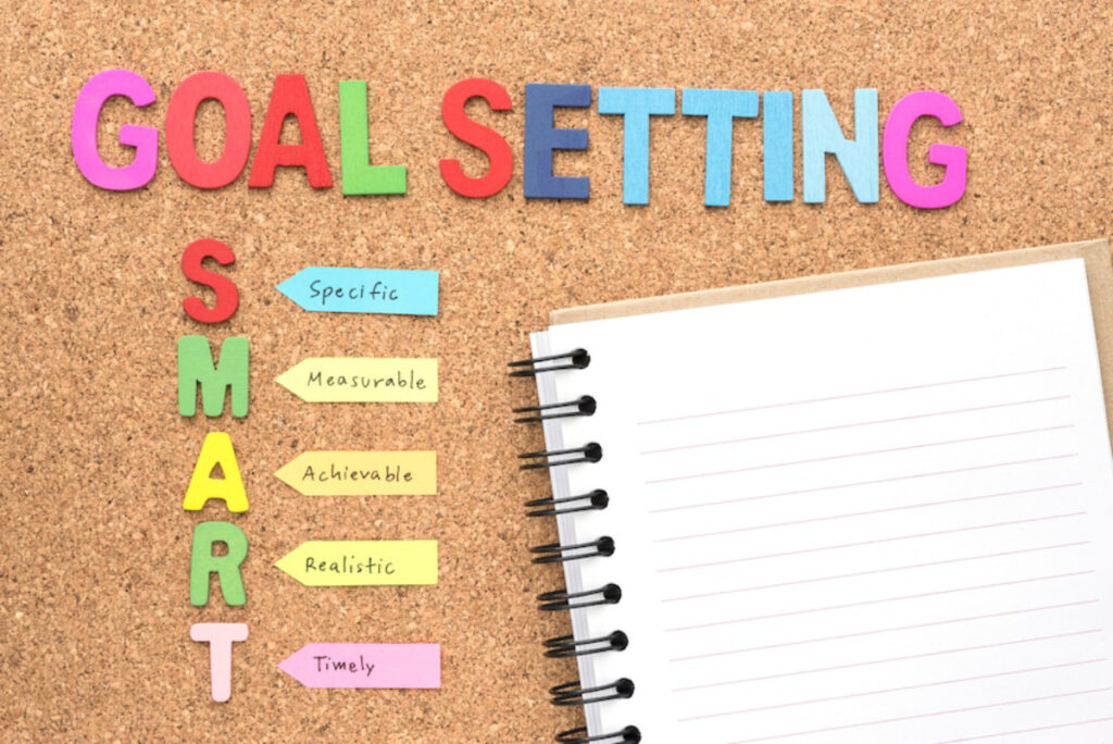Colorful letters spell Goal Setting on a corkboard, accompanied by the SMART criteria: Specific, Measurable, Achievable, Realistic, Timely, and a blank notebook.