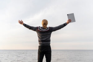 A person with long hair, wearing a patterned sweater, stands by the sea with arms outstretched, holding a tablet against a cloudy sky.