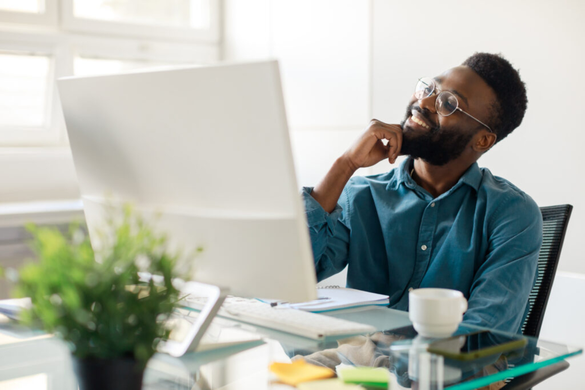 A person in a blue shirt sits at a desk, deep in thought, in a bright office filled with plants and a computer.
