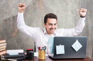 A person in a white shirt celebrates triumphantly at a desk with books, stationery, and a laptop covered in sticky notes.
