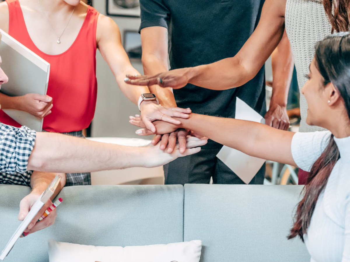 A group of diverse individuals joining hands in a collaborative gesture, surrounded by documents and a casual meeting space.