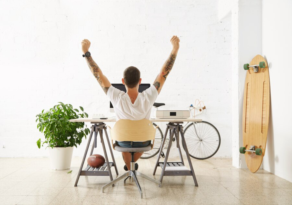 A man with tattoos raises his arms in celebration while sitting at a stylish desk in a bright, modern workspace.