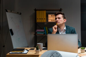A focused individual in a suit sits at a desk with a laptop, surrounded by office supplies and a whiteboard.