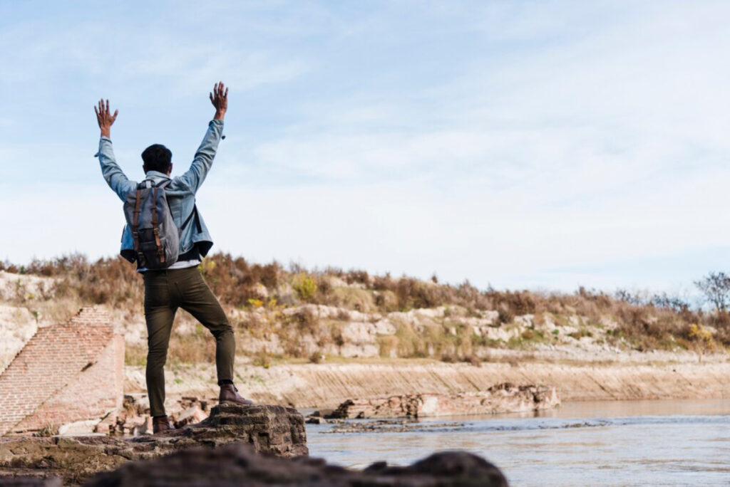 A man stands on a rock with arms raised, celebrating beside a tranquil river against a backdrop of autumn foliage and blue sky.