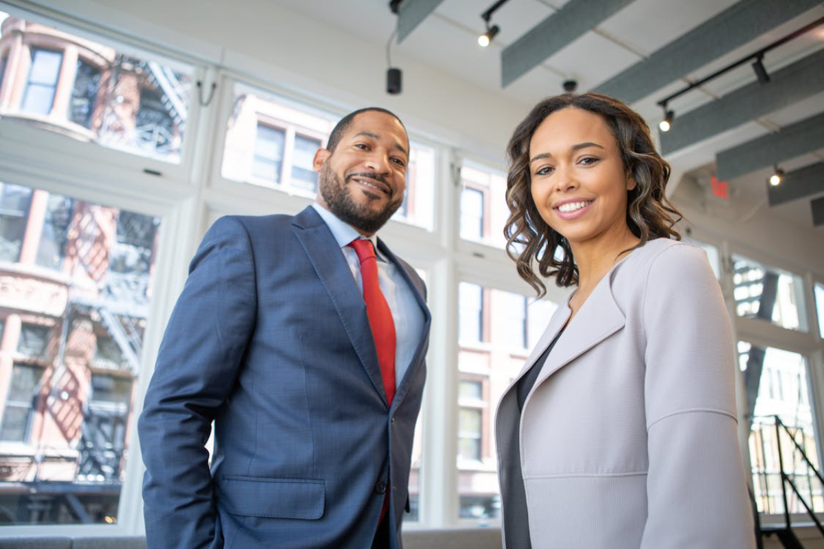 Two professionally dressed individuals stand in a well-lit modern space, with large windows revealing an urban backdrop.