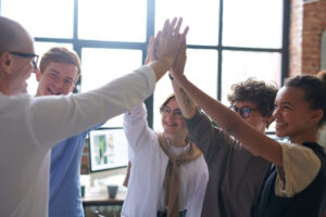 A group of diverse individuals enthusiastically high-fiving each other in a bright, modern workspace with large windows.