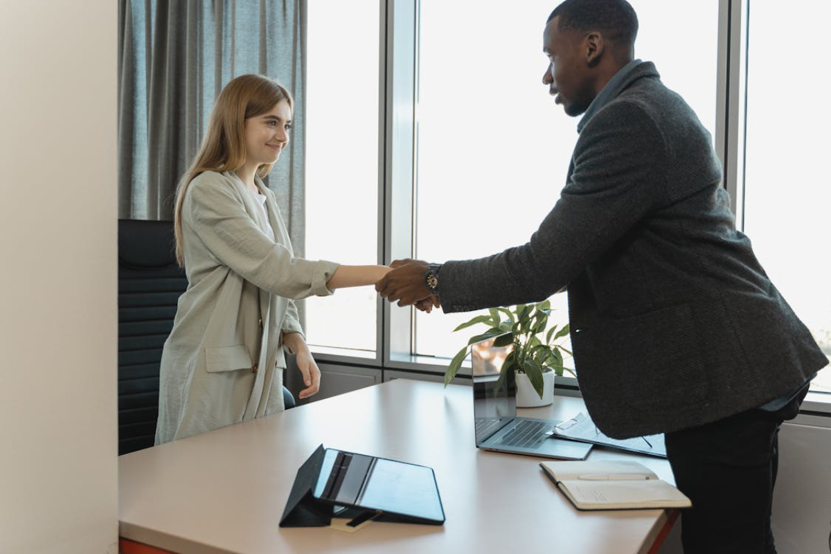 A professional handshake between two people in an office setting, with a laptop and a notebook visible on a desk.
