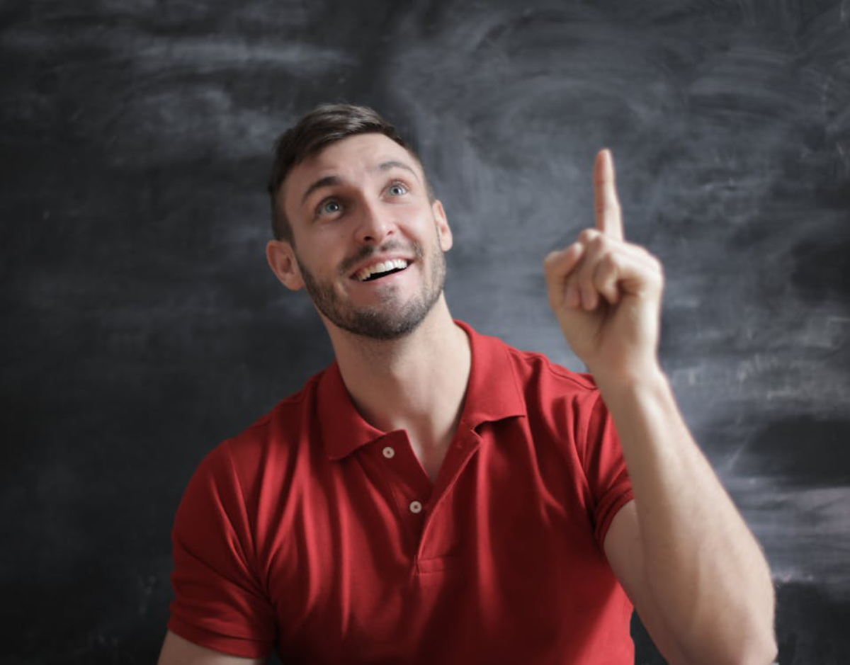 A person in a red polo shirt raises one finger in a thoughtful gesture against a dark chalkboard background.