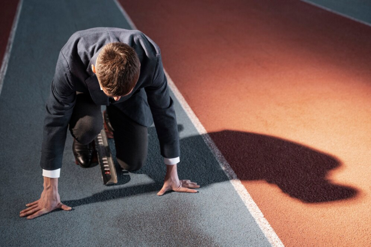 A man in a suit prepares to race on a running track, illustrating the contrast between formal attire and athletic readiness.