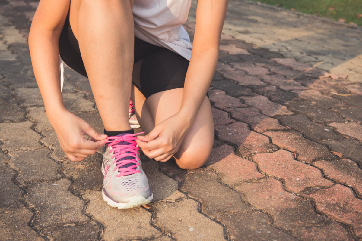 A person kneels on a pathway, tying the pink shoelaces of their grey running shoes, prepared for exercise.