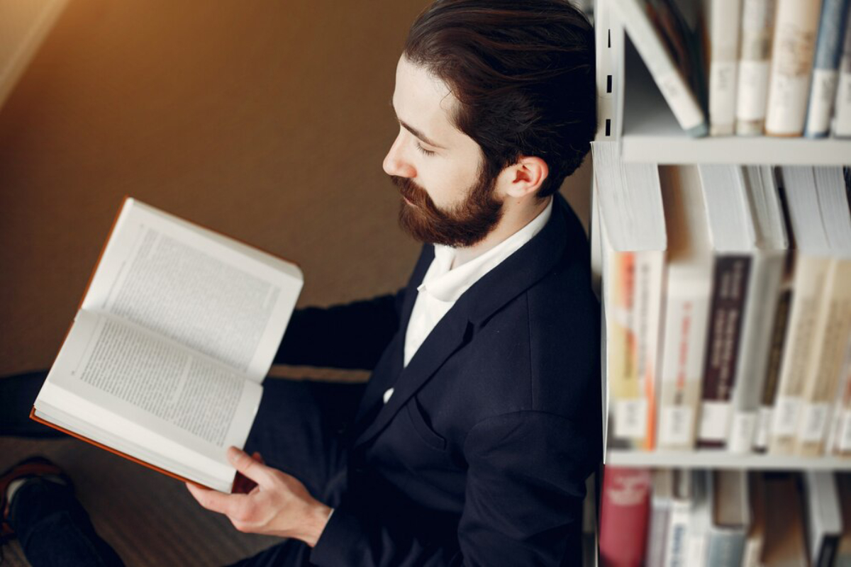 A person in a black blazer sits on the floor, deeply engaged in reading a book, surrounded by library shelves filled with books.