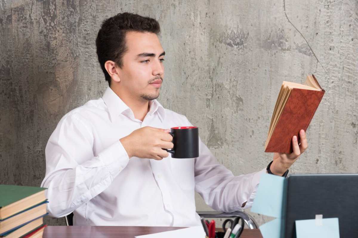 A person in a white shirt holds a mug and a book while seated at a desk surrounded by stacks of books and stationery.