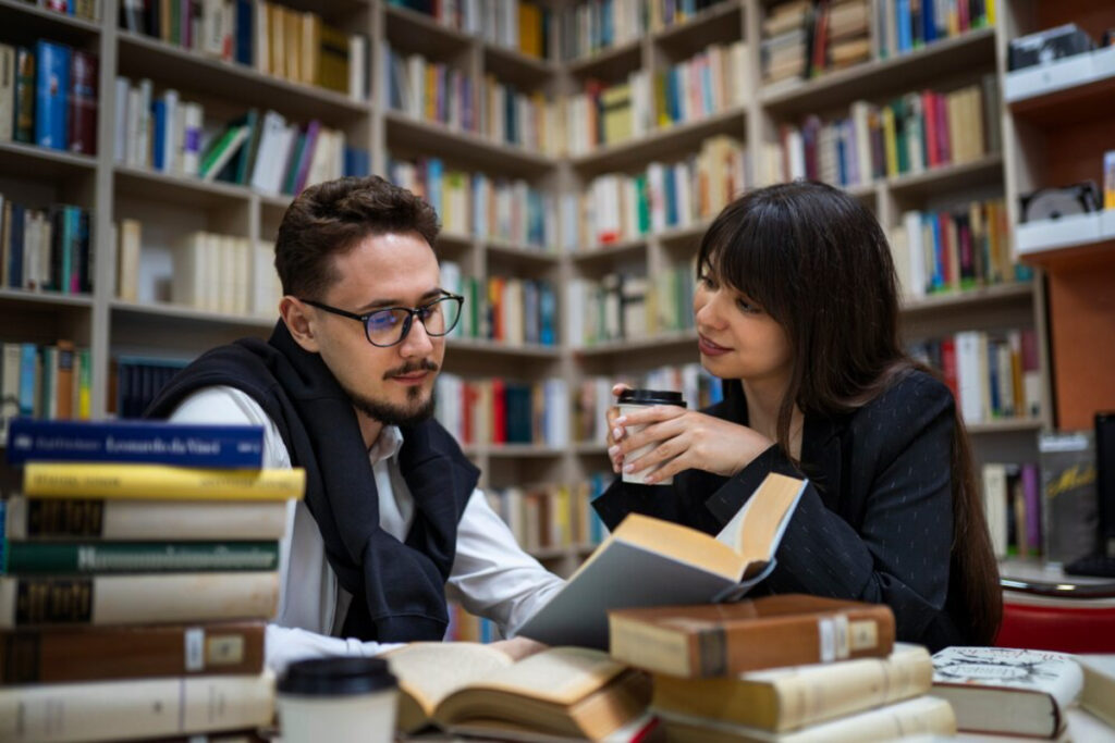 A cozy library setting with two individuals discussing over books, surrounded by a collection of stacked and shelved literature.