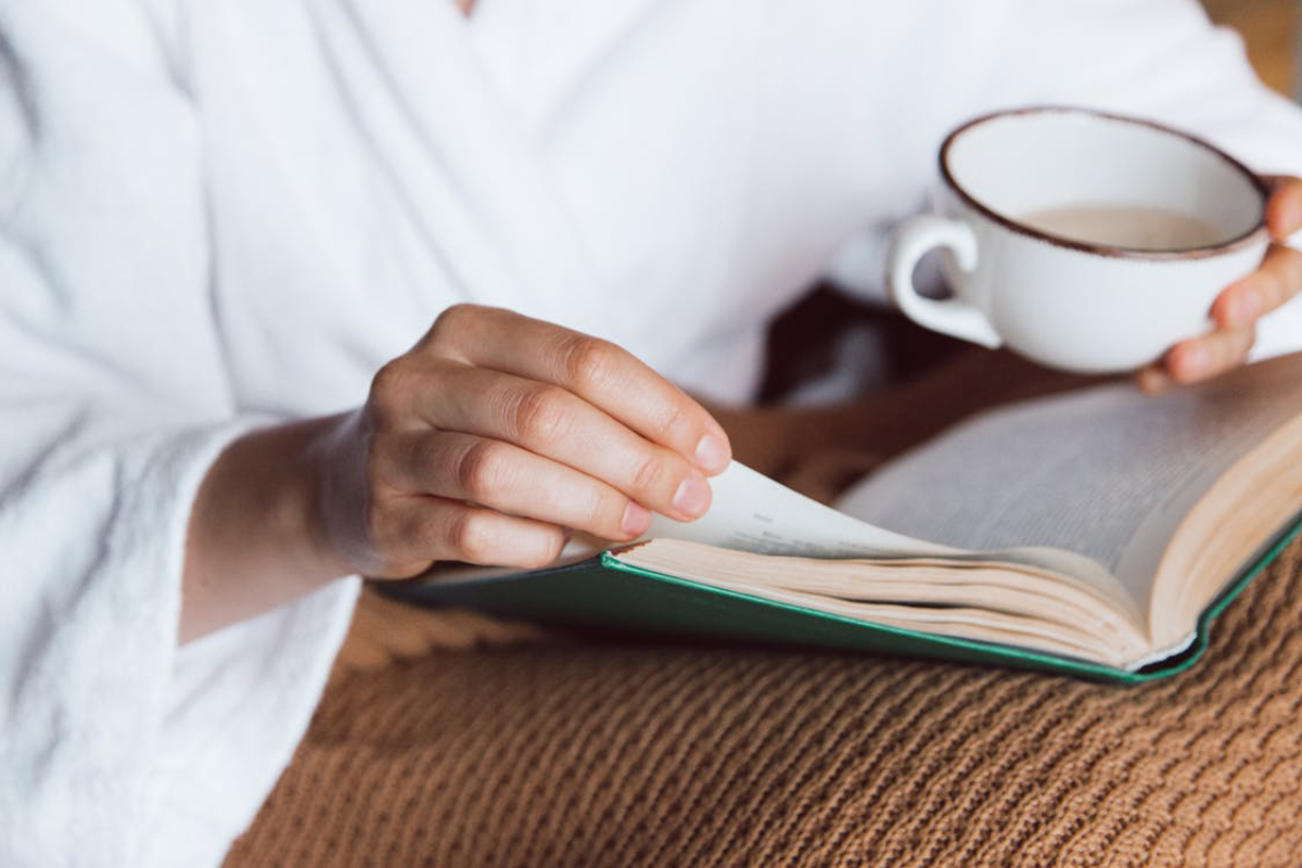 A person in a white robe enjoys reading a book while holding a cup of coffee, seated on a cozy, textured blanket.