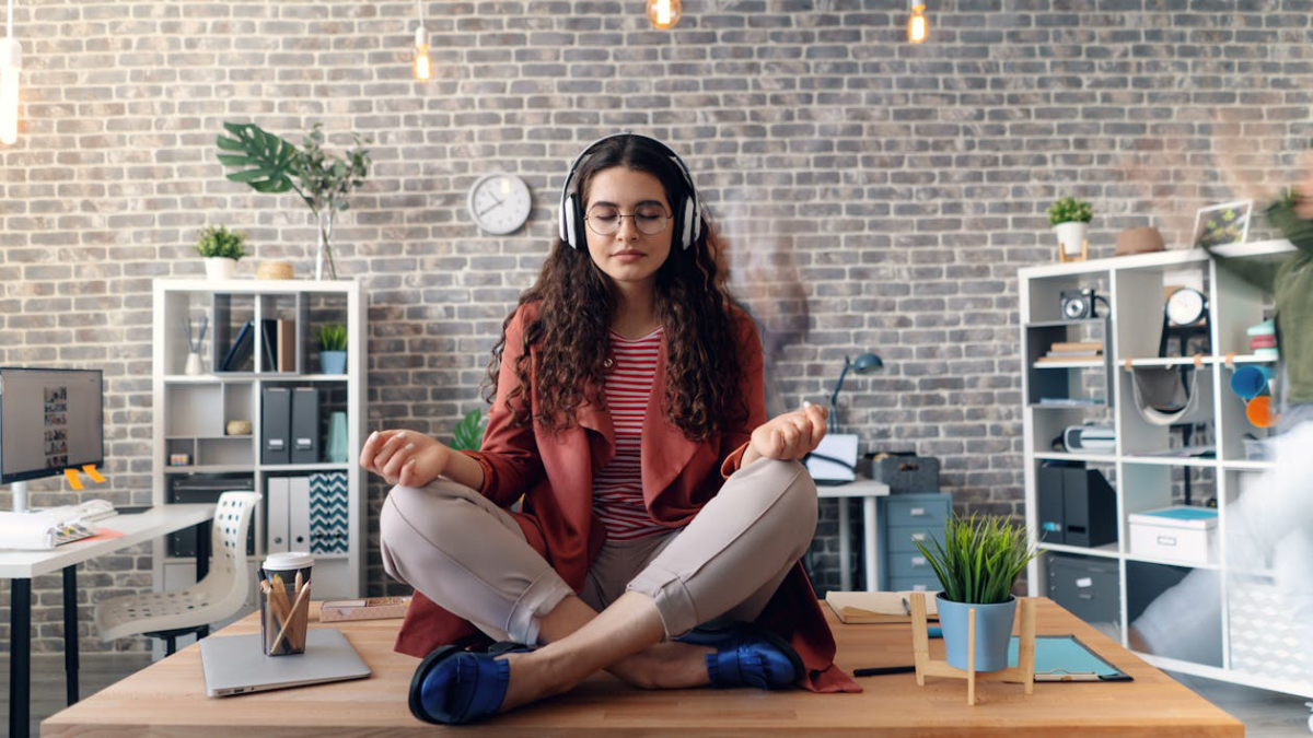 A woman in a red jacket meditates on a desk in a modern office, wearing headphones.