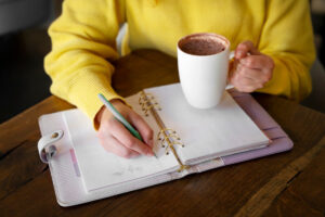 A person wearing a yellow sweater writes in a planner while holding a white mug of hot drink, seated at a wooden table.