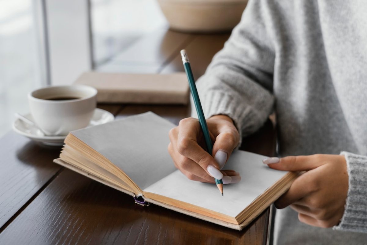 A person in a cozy sweater writes in an open notebook with a pencil, while a steaming cup of coffee sits nearby.