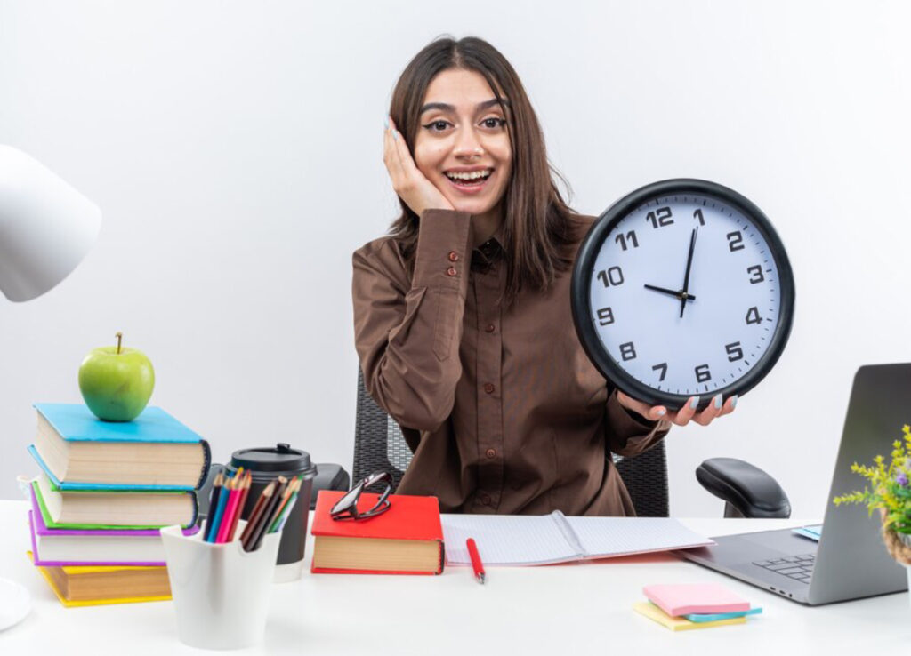 A person sits at a desk cluttered with books, stationery, and a laptop, holding a clock, signaling stress or urgency.