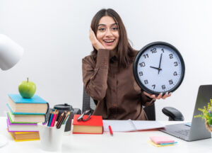 A person sits at a desk cluttered with books, stationery, and a laptop, holding a clock, signaling stress or urgency.