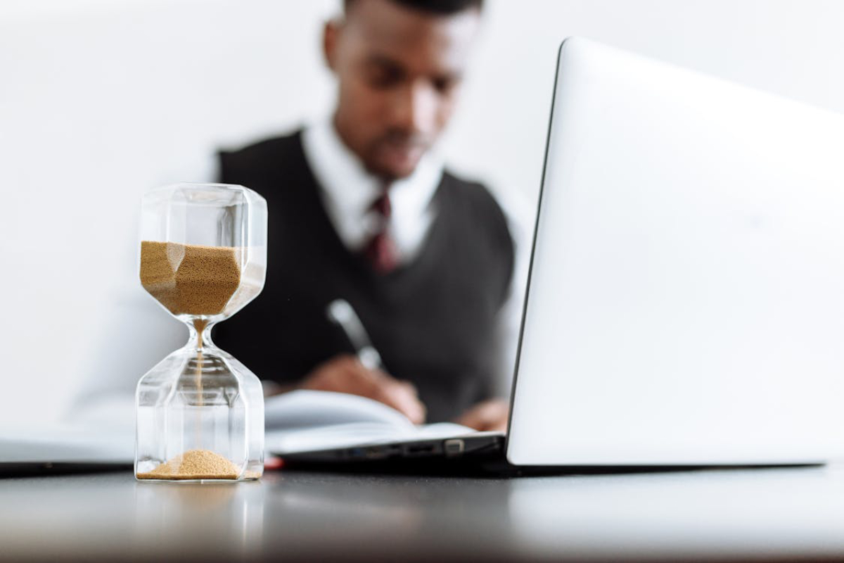 A glass hourglass filled with sand sits on a desk next to a laptop, with a person writing in a notebook in the background.