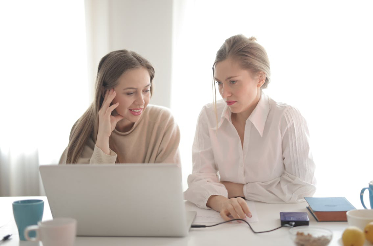 Two women are seated at a table with a laptop, engaged in discussion. Coffee cups and a notebook are visible on the table.