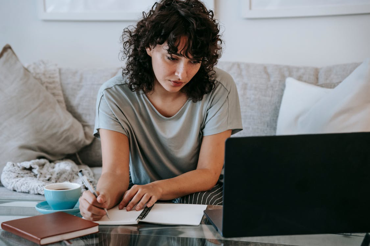 A person with curly hair sits on a couch, writing in a notebook with a cup of coffee and a laptop in front of them.