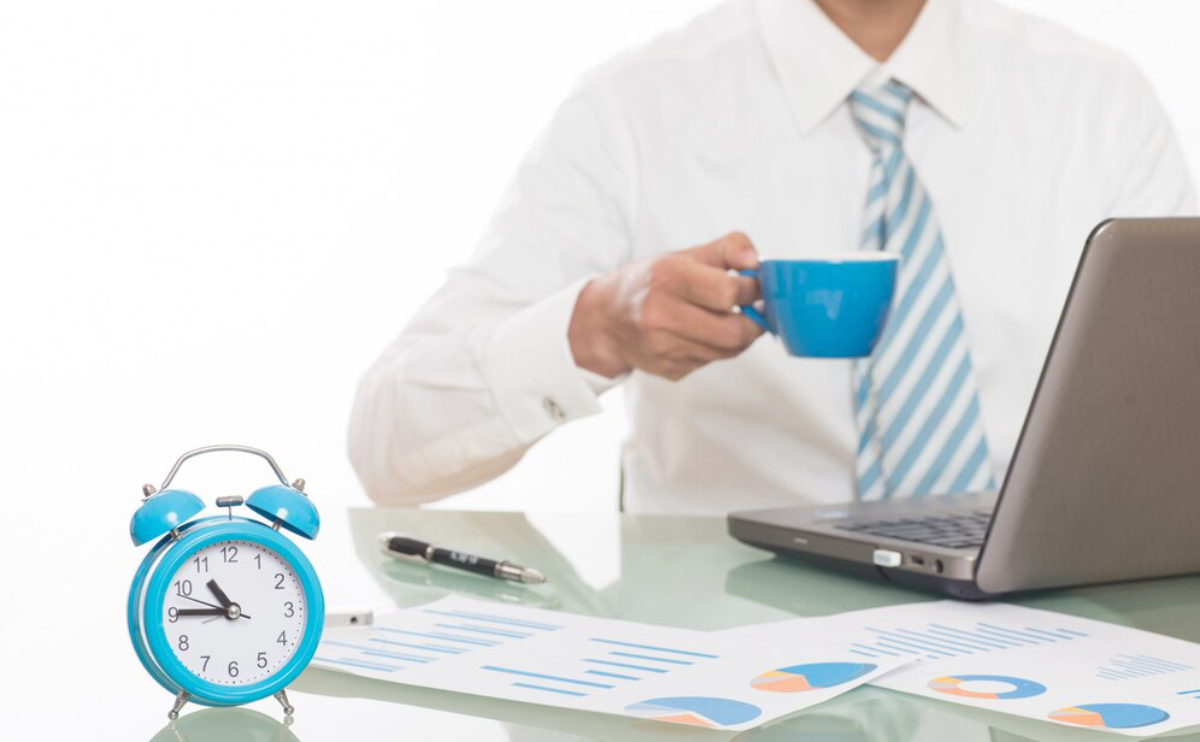 A businessman in a white shirt and tie holds a blue cup while seated at a desk with a laptop and a blue alarm clock.
