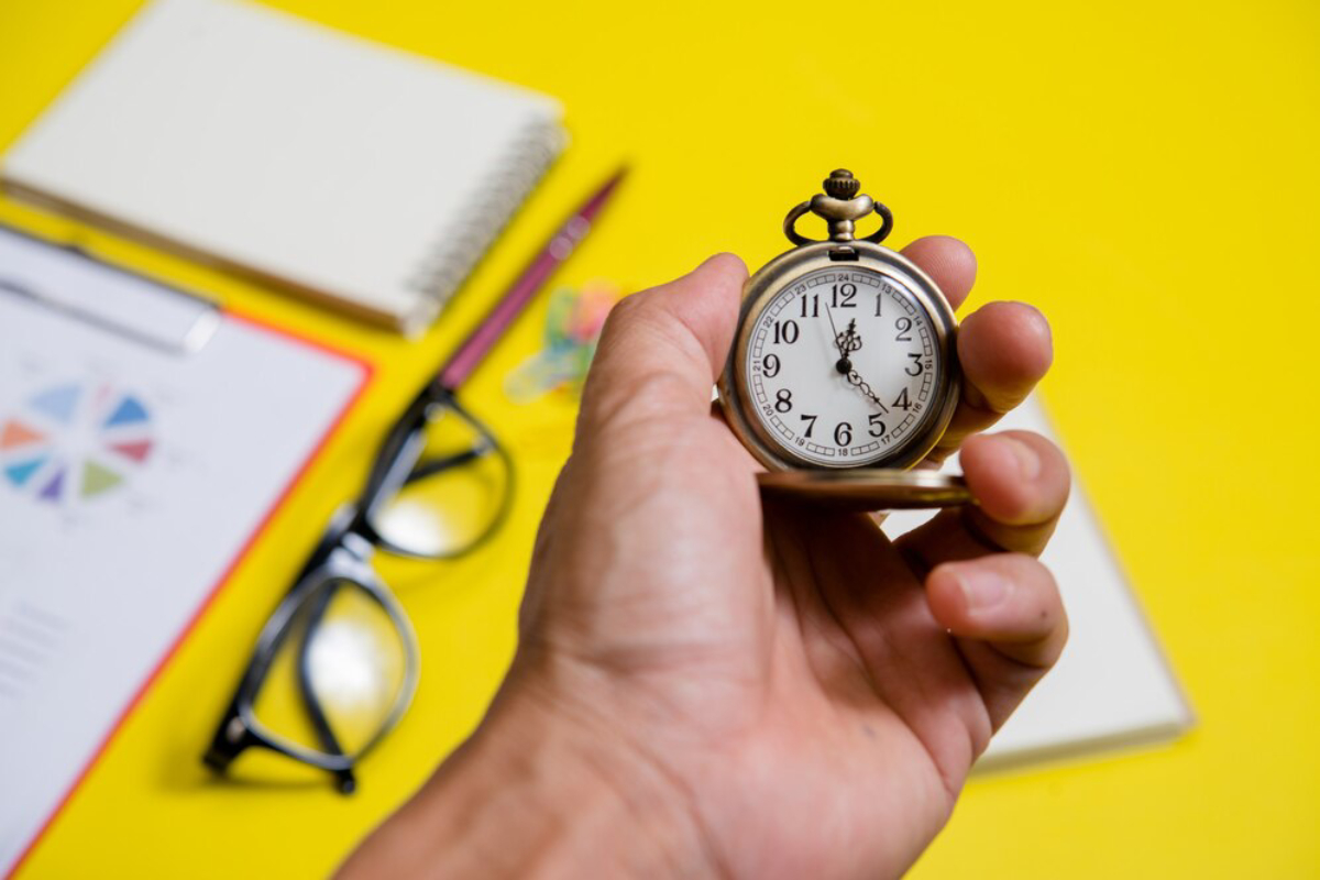 A hand holding a vintage pocket watch over a bright yellow surface with a notepad, glasses, and stationery in the background.