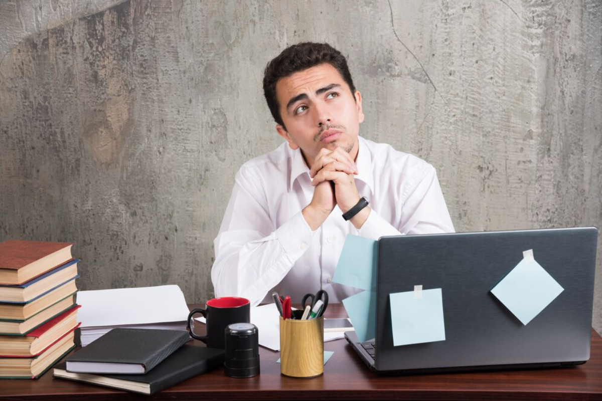 A person sits at a desk with a laptop, surrounded by books, a coffee mug, and stationery, against a concrete wall backdrop.