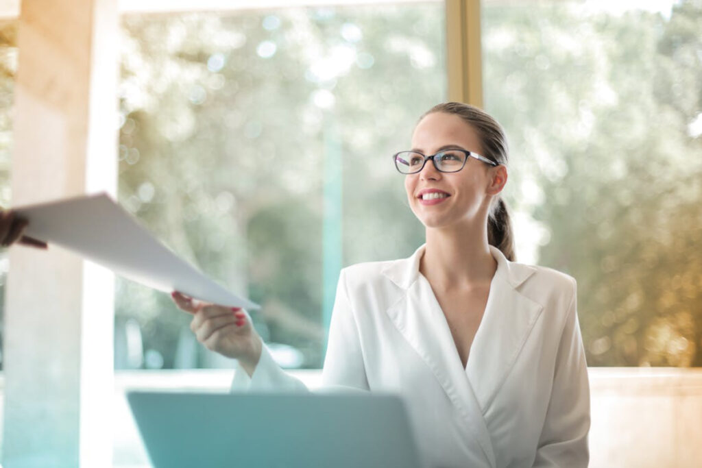 A woman in a white blazer receives a document from a person outside the frame, inside a bright office with a blurred background.